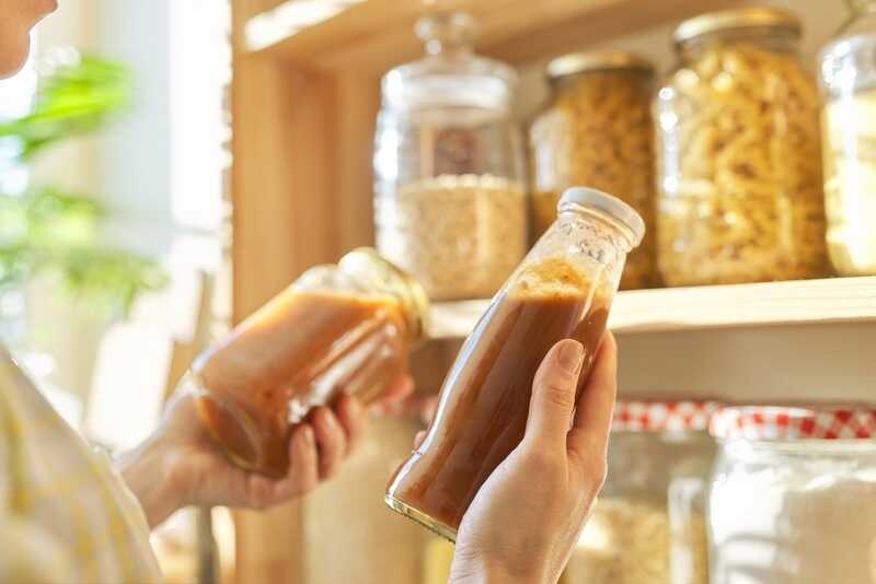 Woman holding bottle with ketchup, picking food from storage cabinet in kitchen, storage with wooden shelves