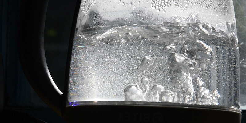 Defocused bubbles in hot water boiling inside glass teapot in bright sunlight. Closeup of boiling water in old transparent electric kettle on kitchen. 