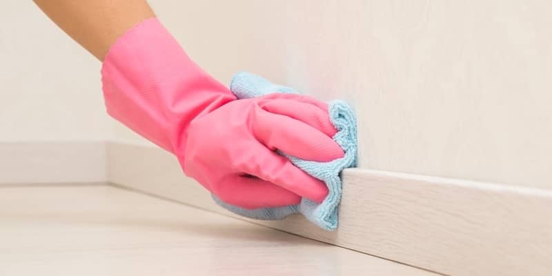 Young adult woman hand in pink rubber protective glove using blue dry rag and wiping light wooden skirting board in room at home