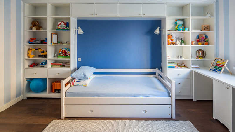 Kid's room in a modern style with striped blue-white walls and a parquet with a carpet on the floor. There is a white bed with pillows, lockers and shelves with toys and books, table.