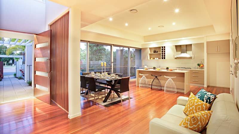 Kitchen with appliances, wooden floor, expensive furnitures can see in this room, door and shiny floor have made with wood, pillows with designs on the sofa, green trees, grand front entrance.