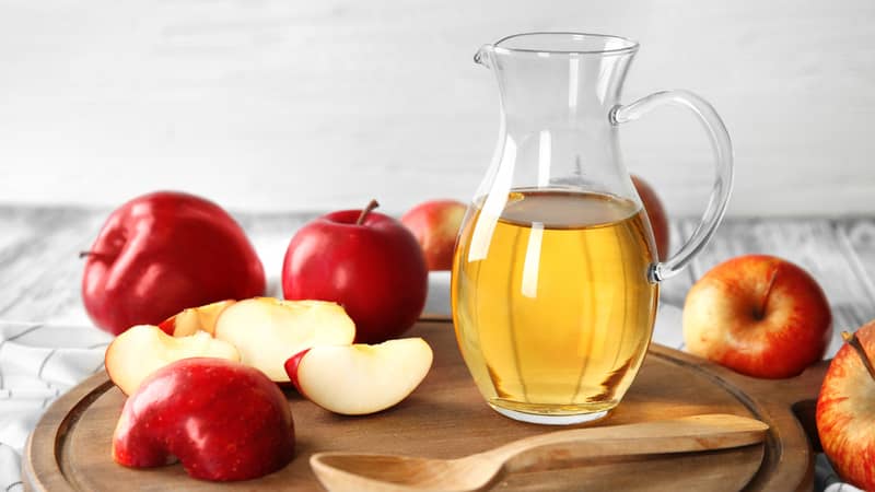 Glass jug with apple vinegar and fresh fruit on wooden board.