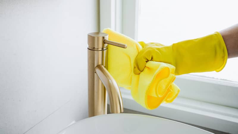 female hand in yellow gloves wiping the faucet in the sink with cloth