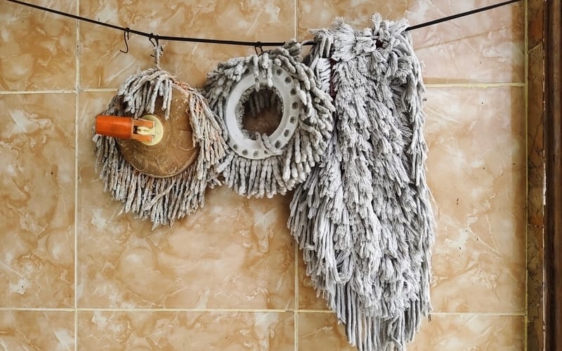 Various used mop heads hanging to dry against a beige tiled wall,