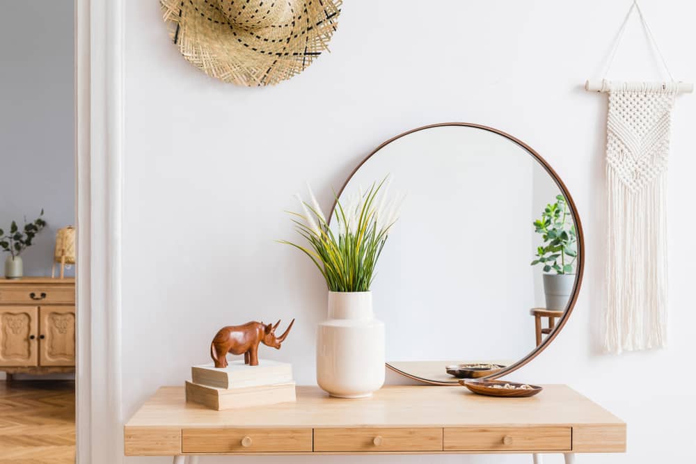 Sunny boho interiors of apartment with mirror, dressing table, furnitures, flowers, plants, rattan hat, sculpture, macrame and design accessories.