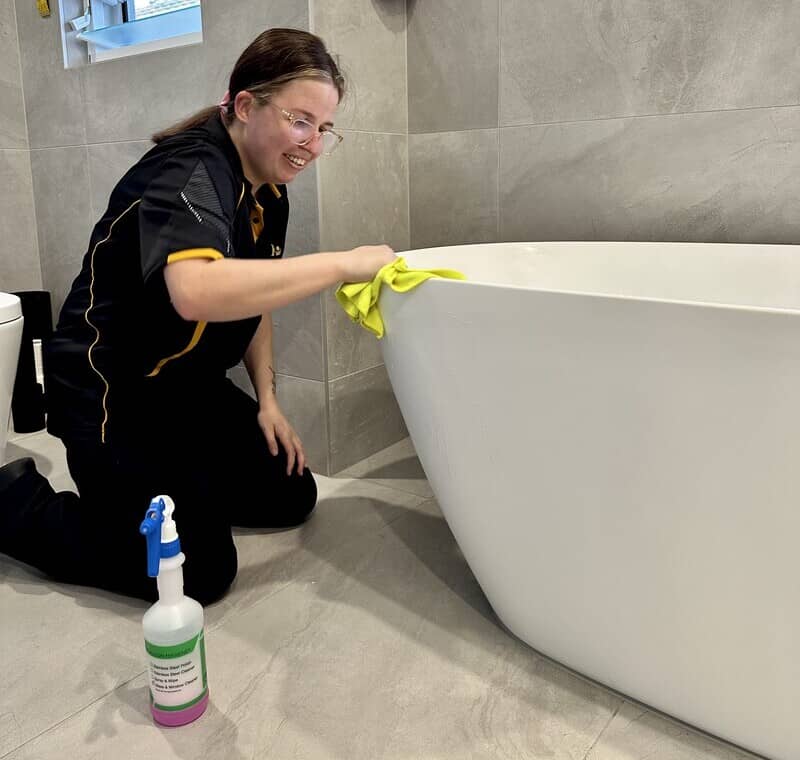 Kirsten wiping the side of the tub with a microfibre cloth and a cleaning solution set beside her