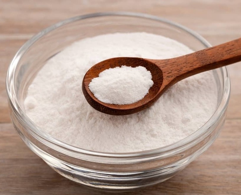 Scooping baking powder with a wooden spoon from a clear glass bowl.