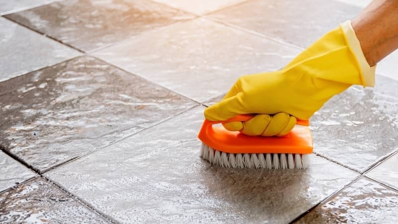 Hands wearing yellow rubber gloves are using a nylon brush to scrub the tile floor with a floor cleaner.