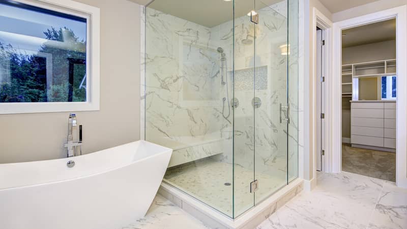 Sleek bathroom features freestanding bathtub atop marble floor placed in front of glass shower accented with rain shower head and gray and white marble surround.