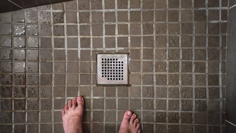 Looking down to shower drain grid, wet tiles on floor, man feet visible in lower part