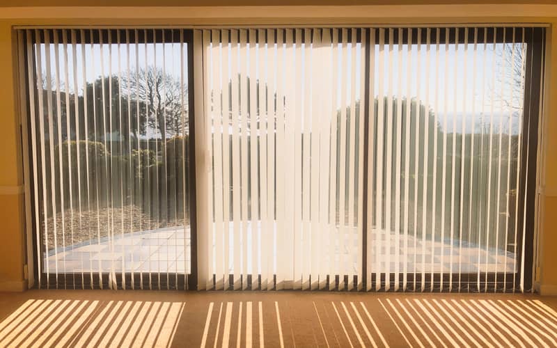 sunlight shining through full length white vertical blinds in front of three glass sliding French doors 