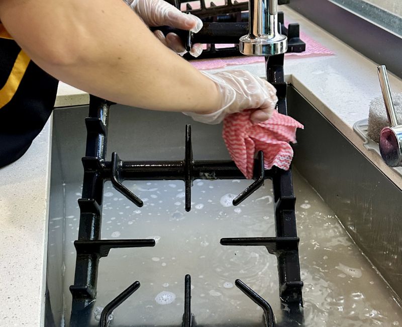 Maid2Match cleaner cleaning stove top grates in the sink with soapy water and microfibre cloth