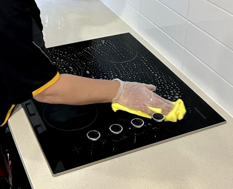 Maid2Match cleaner wiping a glass cooktop with a microfibre cloth in a white tiled kitchen.