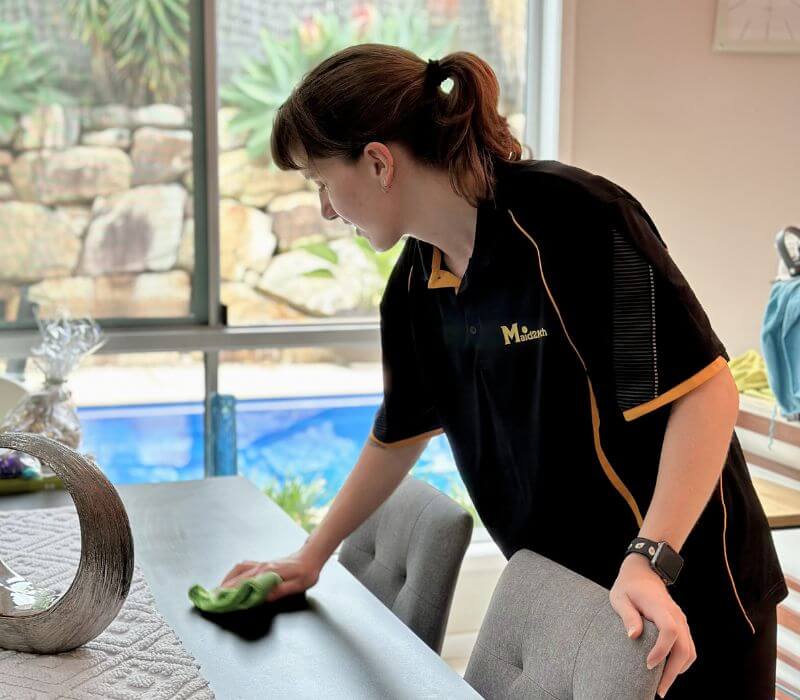 Maid2Match Brisbane experienced and friendly house cleaner Kayleigh cleaning a dining table in the living room with microfibre cloth