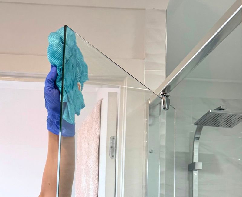 Hand wearing gloves cleaning shower glass with bleach and microfibre towel