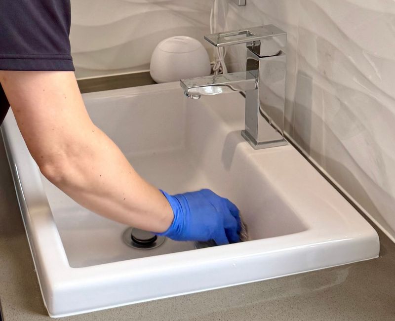 Hand wearing protective gloves cleaning bathroom sink with bleach